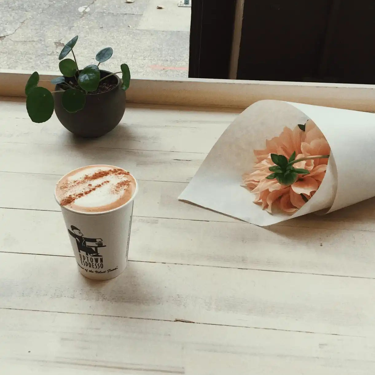 Coffee in a to-go cup, potted plant, and pink flower bouquet on a light wood table by a window.