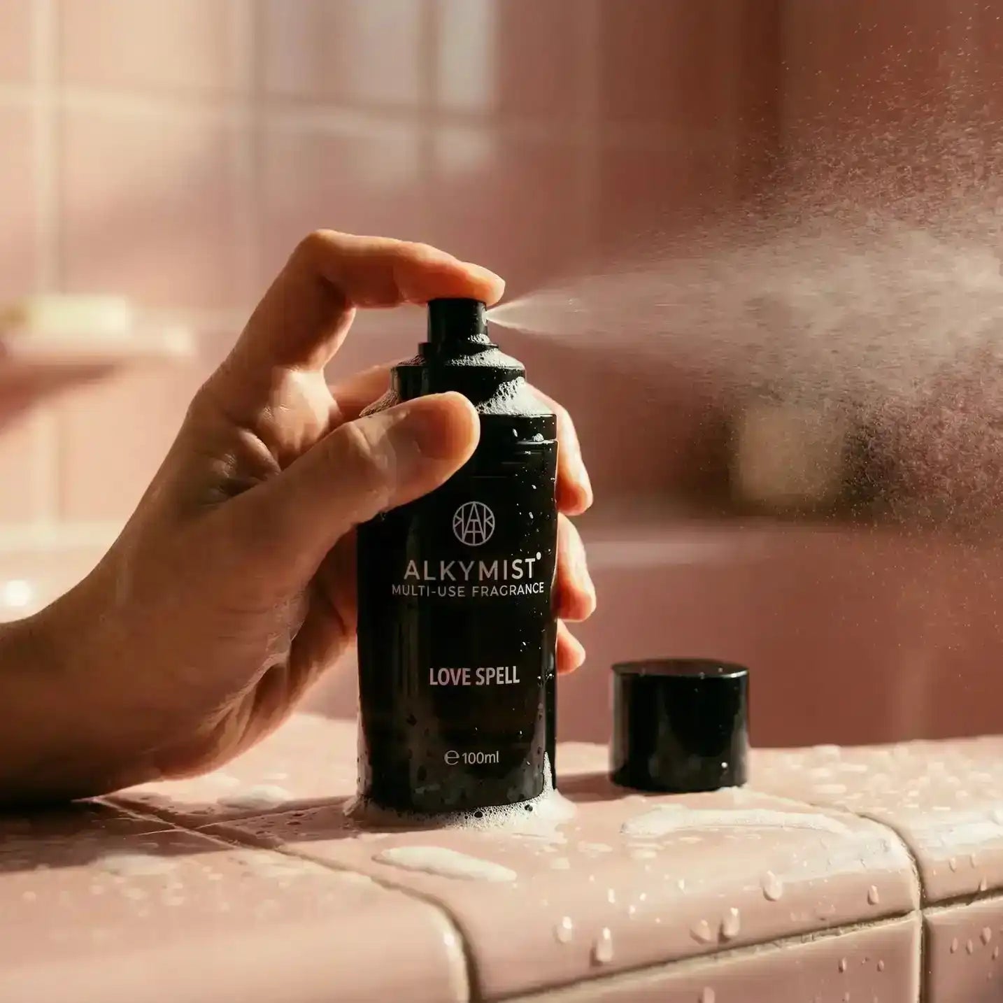 Hand spraying AEMBR ALKYMIST multi-use fragrance bottle 'Love Spell' on pink bathroom tiles