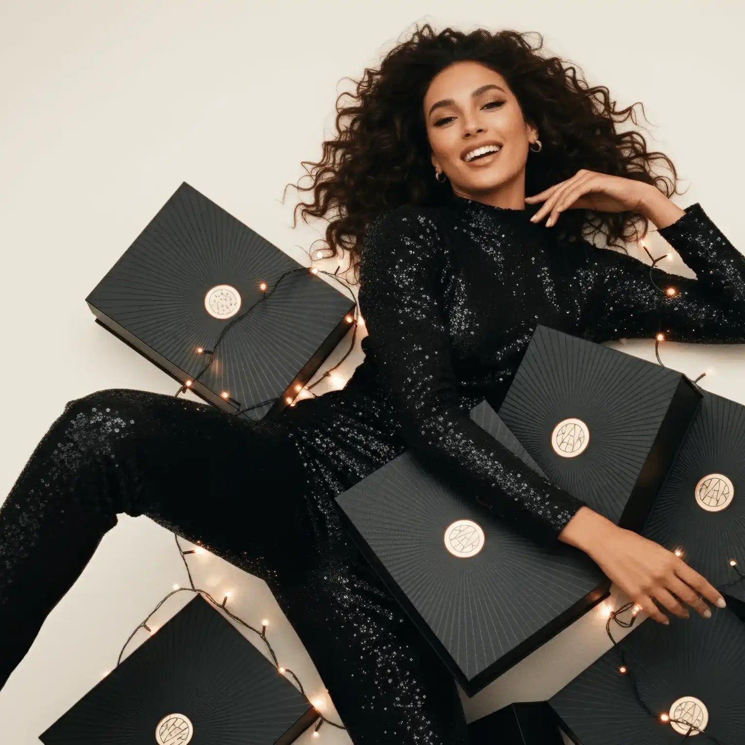 Woman in black sequin outfit smiling, surrounded by AEMBR gift boxes and string lights.