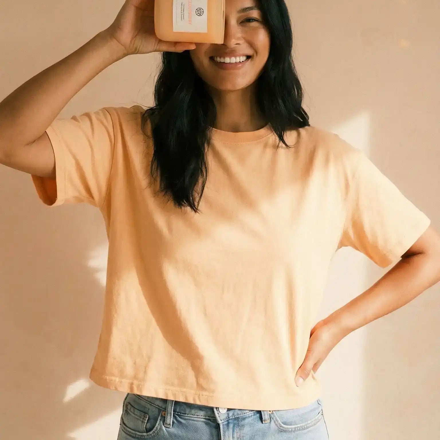 Smiling woman in peach shirt holding AEMBR non-toxic candle in natural light