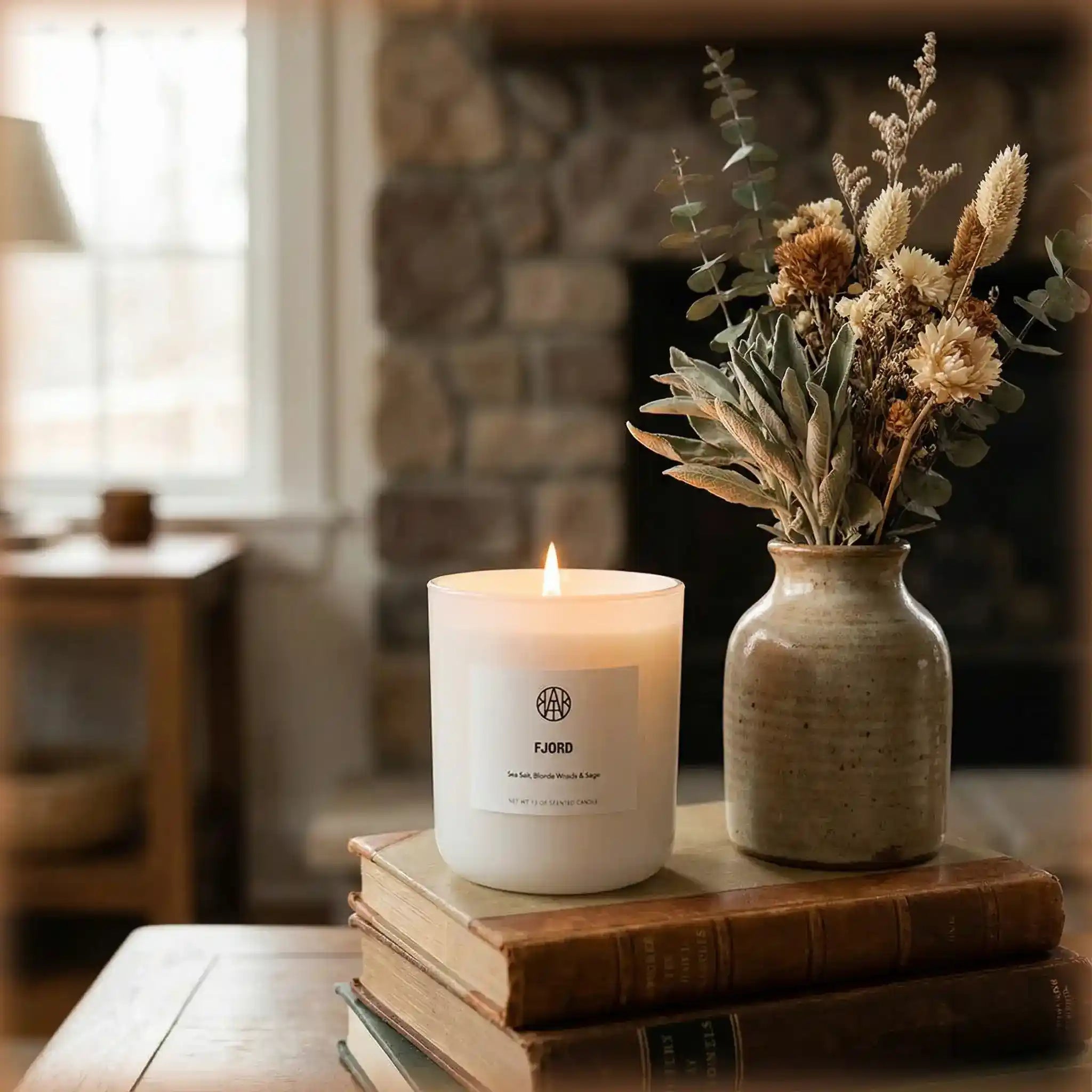 Non-toxic candle burning on stacked books beside a ceramic vase with dried flowers in cozy living room