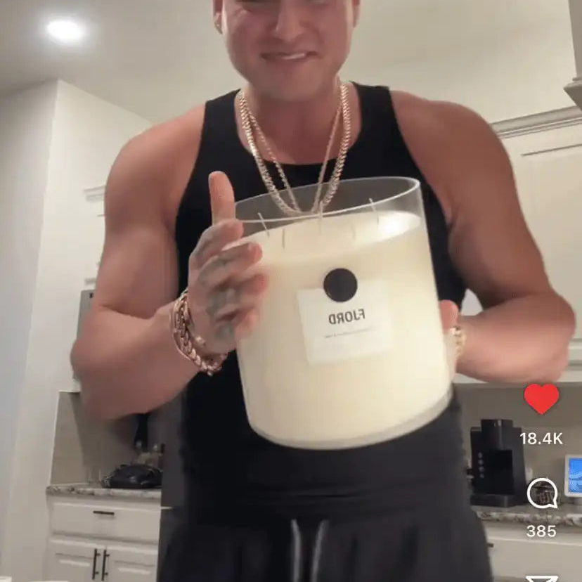 Smiling man in a black tank top holding a large AEMBR FLORD candle in a modern kitchen.