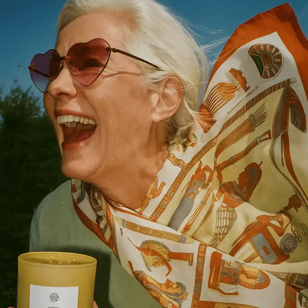 Smiling woman in heart sunglasses with silk scarf holding yellow AEMBR candle outdoors