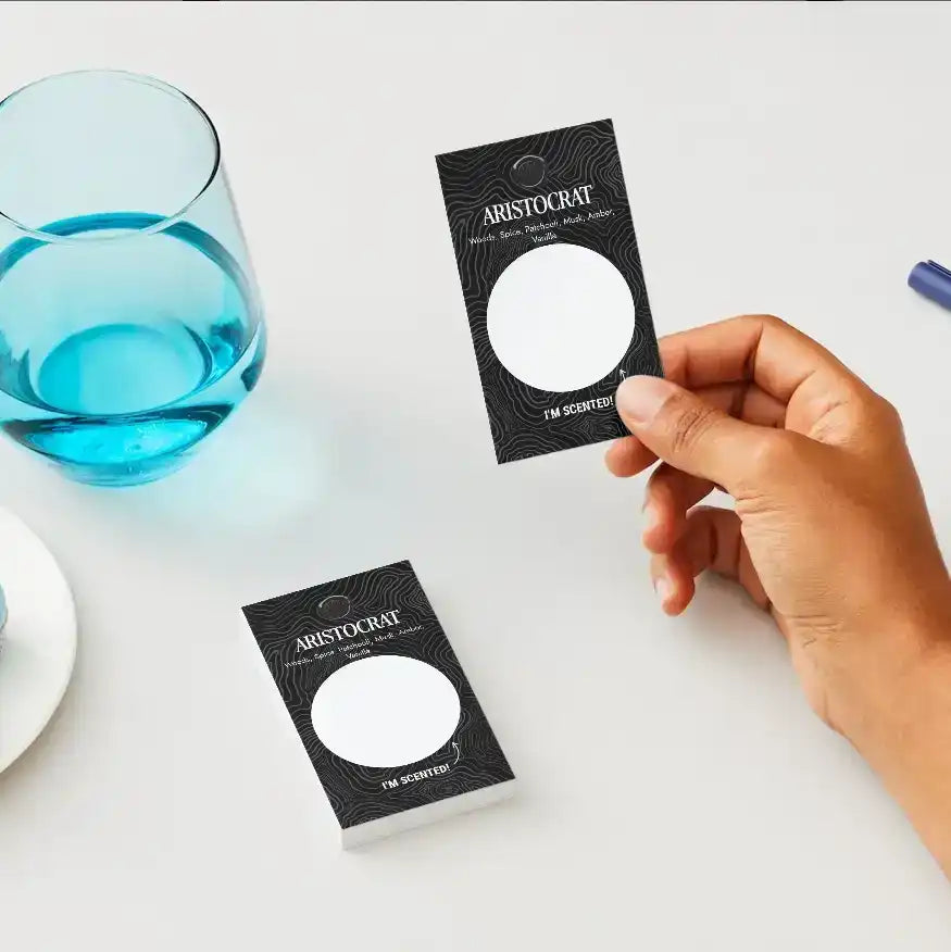 Hand holding AEMBR Aristocrat scented card sample next to glass of water on table