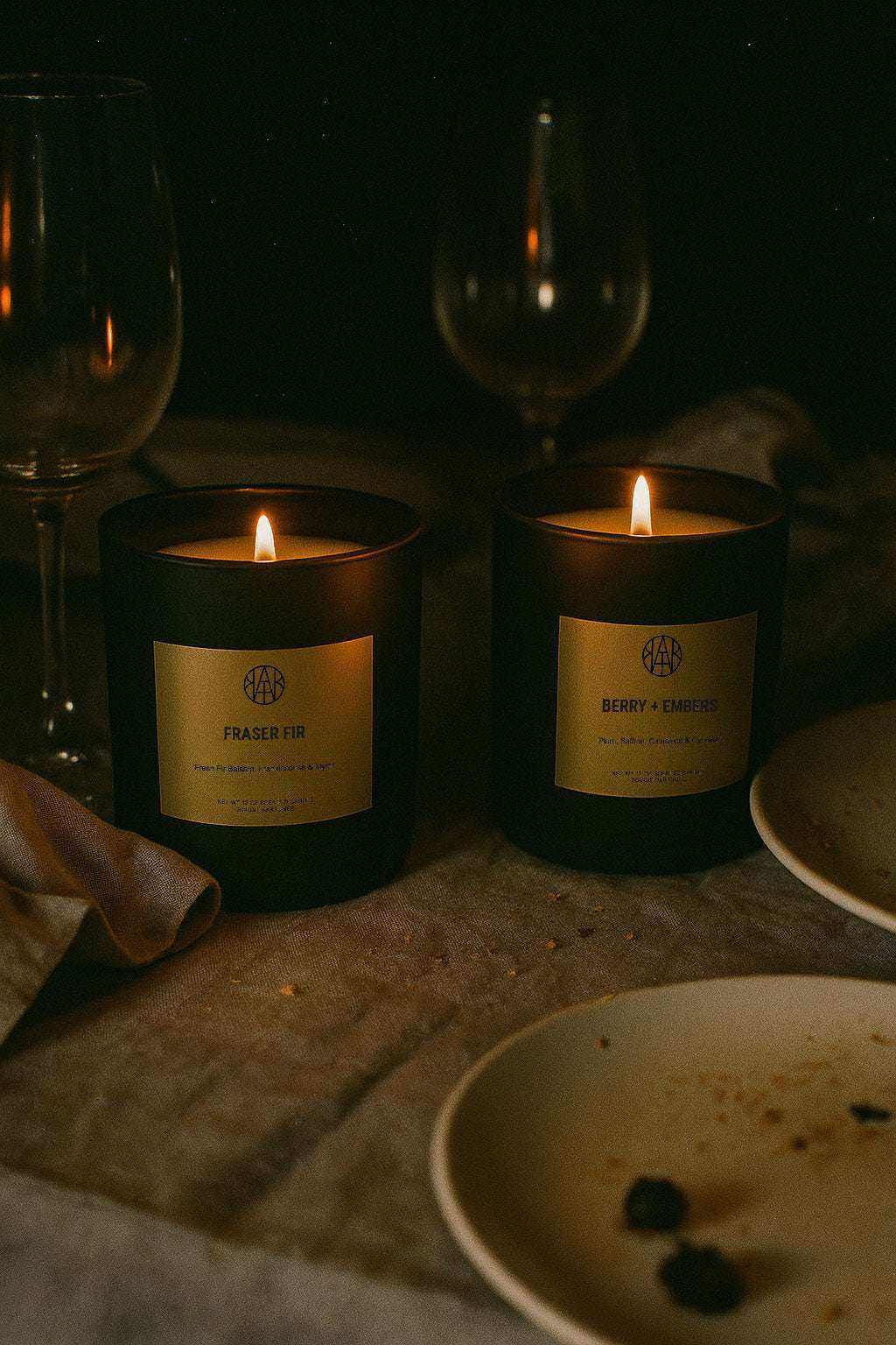 Two lit non-toxic candles on a dinner table, labeled Fraser Fir and Berry + Embers, moody ambiance