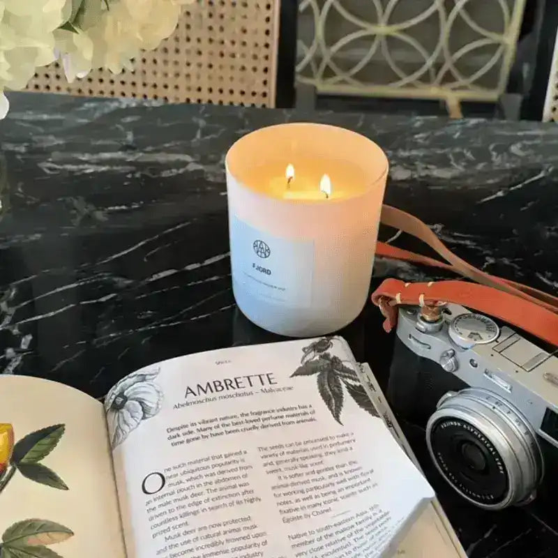 Non-toxic scented candle burning on a black marble table with a camera and open botanical book