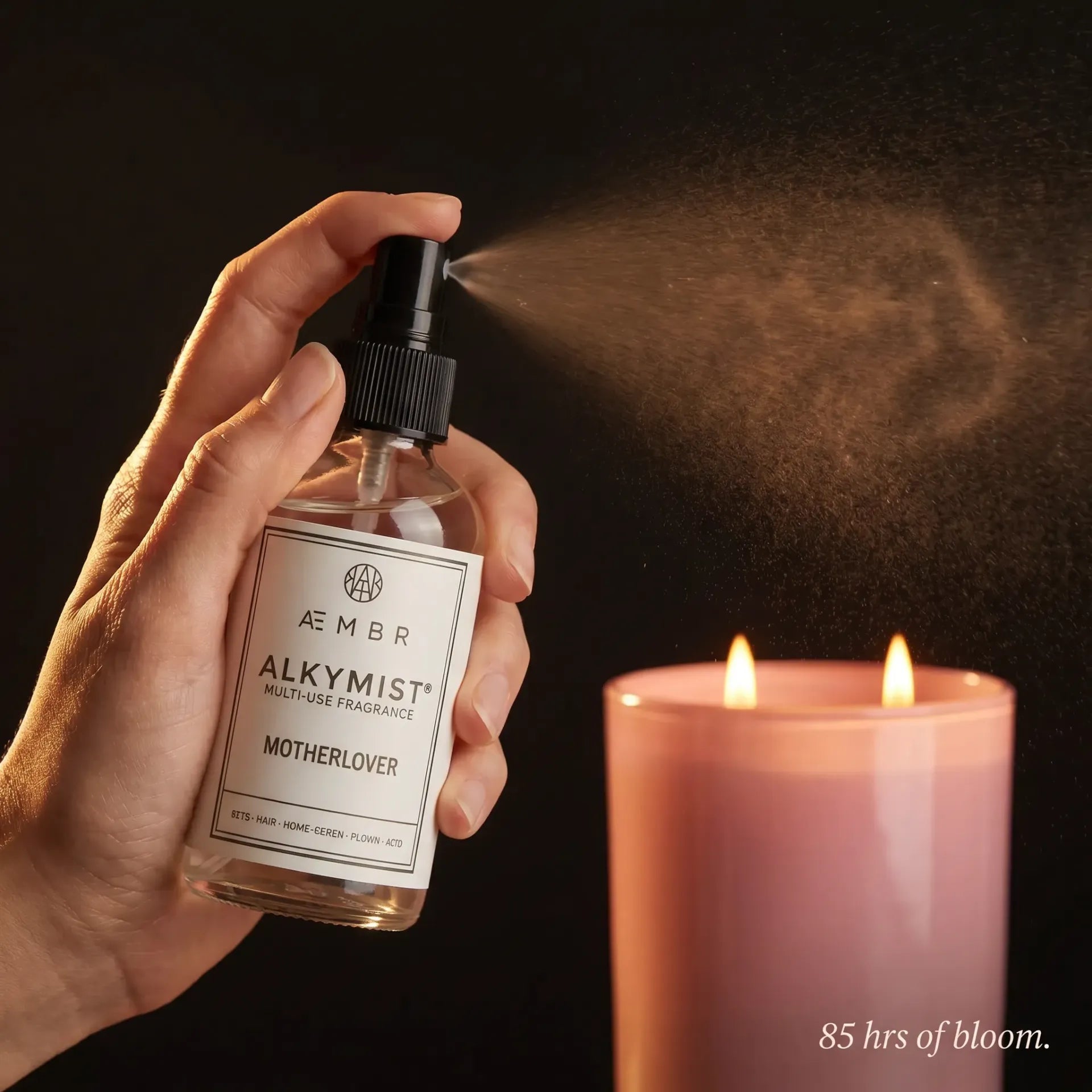 Motherlover Scent Set in use — ALKYMIST Multi-Use Fragrance mid-spray mist with Motherlover candle glowing with two lit wicks — 85 hrs of bloom