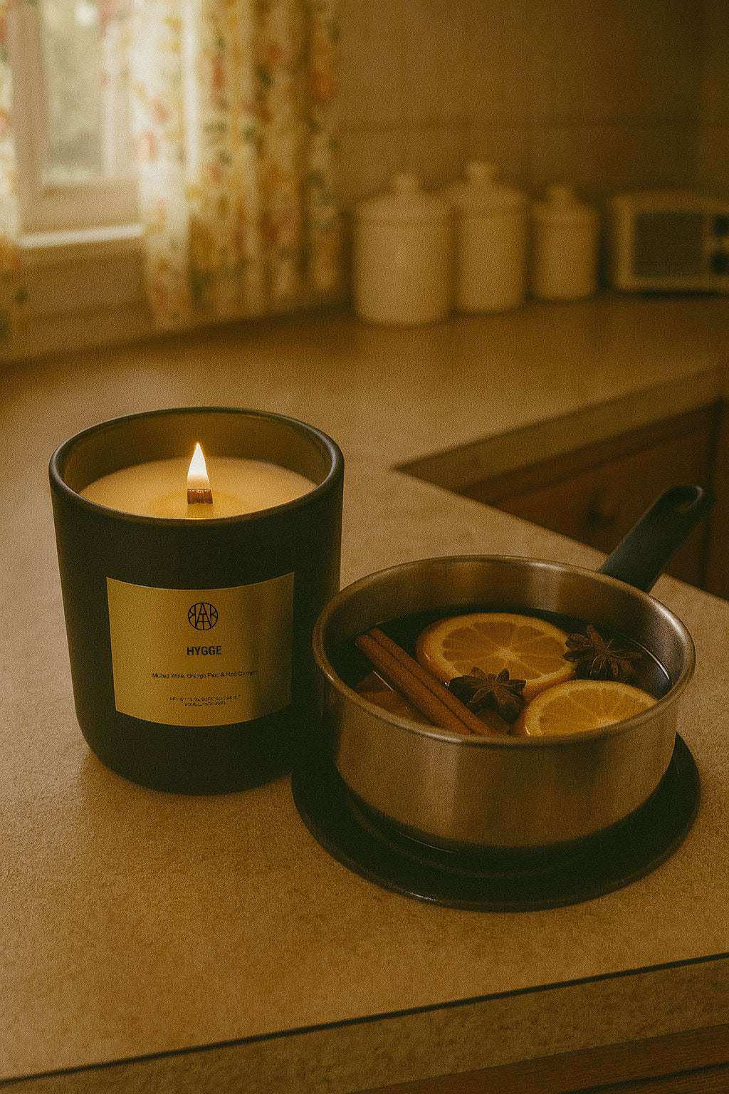 AEMBR Hygge candle burning beside a pan of mulled wine with orange slices and spices on a cozy kitchen counter.
