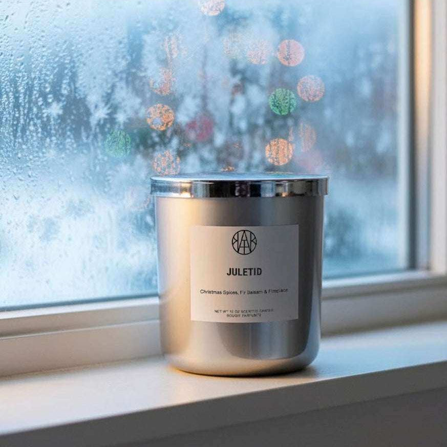 AEMBR Juletid silver candle on a windowsill with frosty glass and festive lights in background.