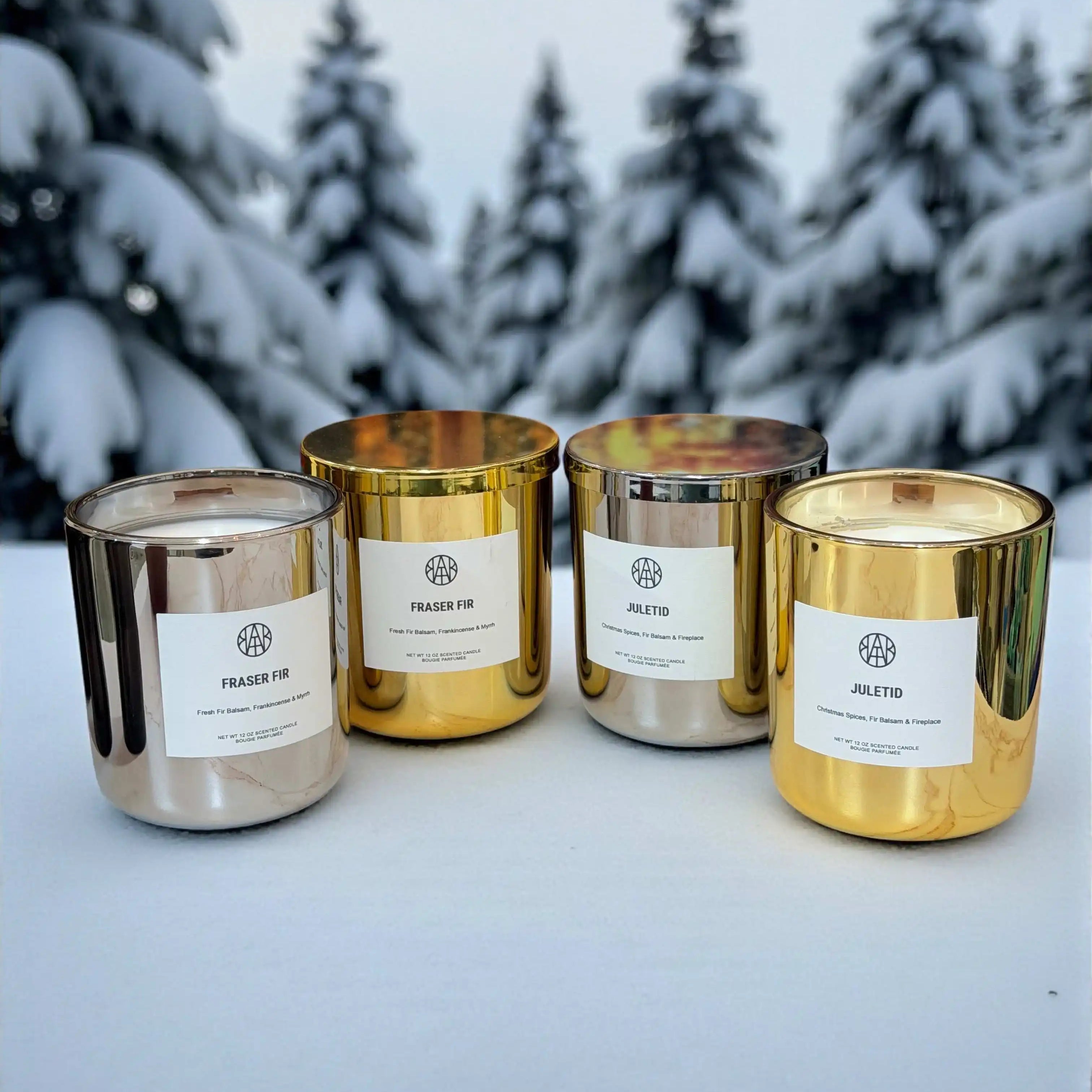AEMBR gold and silver non-toxic candles on snow with snowy pine trees in background