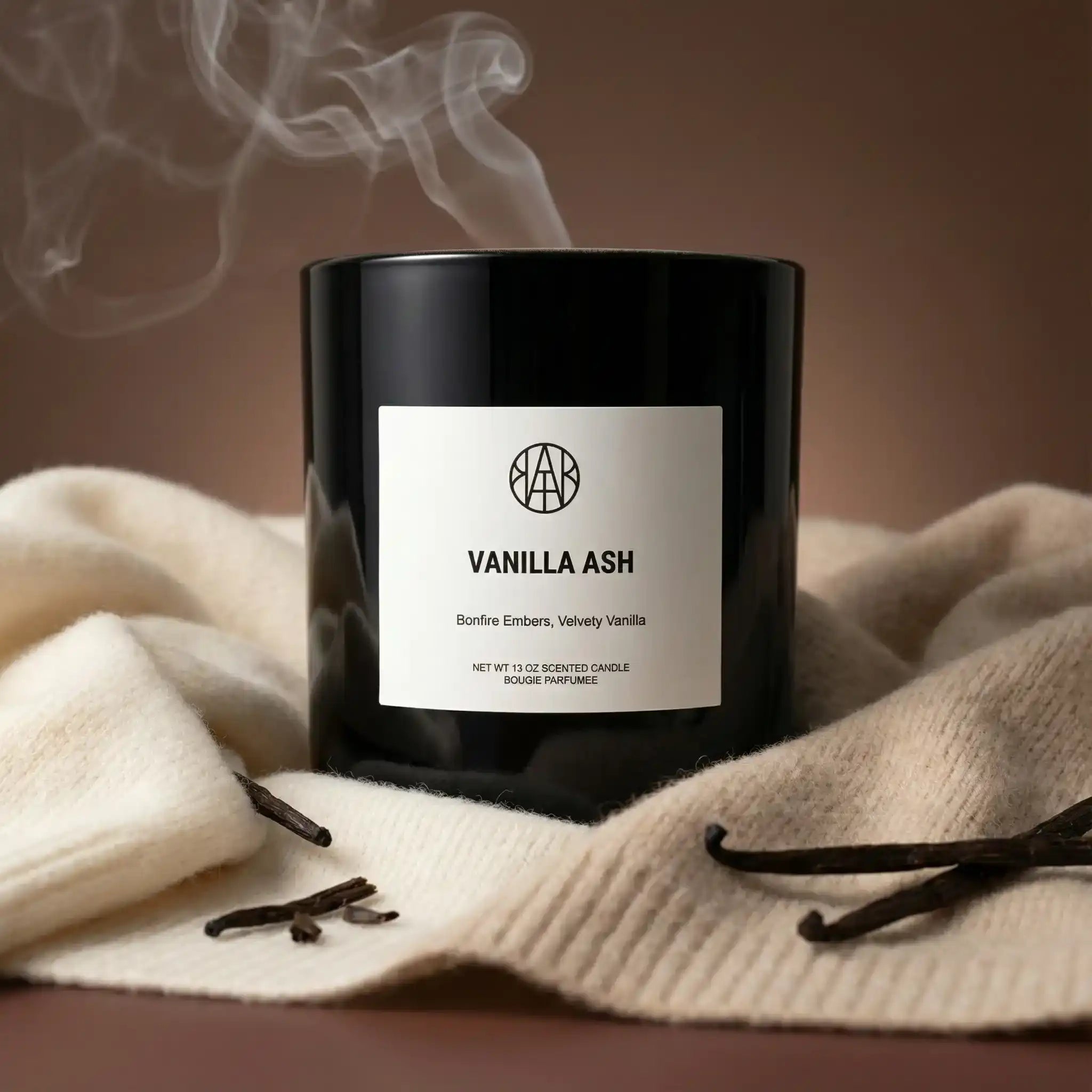 AEMBR Vanilla Ash candle in black glass jar on cozy cream fabric with vanilla pods, smoke rising