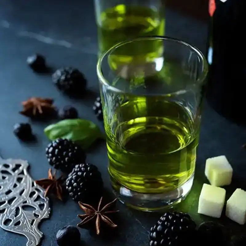 Glass of green liquid with blackberries, star anise, sugar cubes, and herbs on dark tabletop