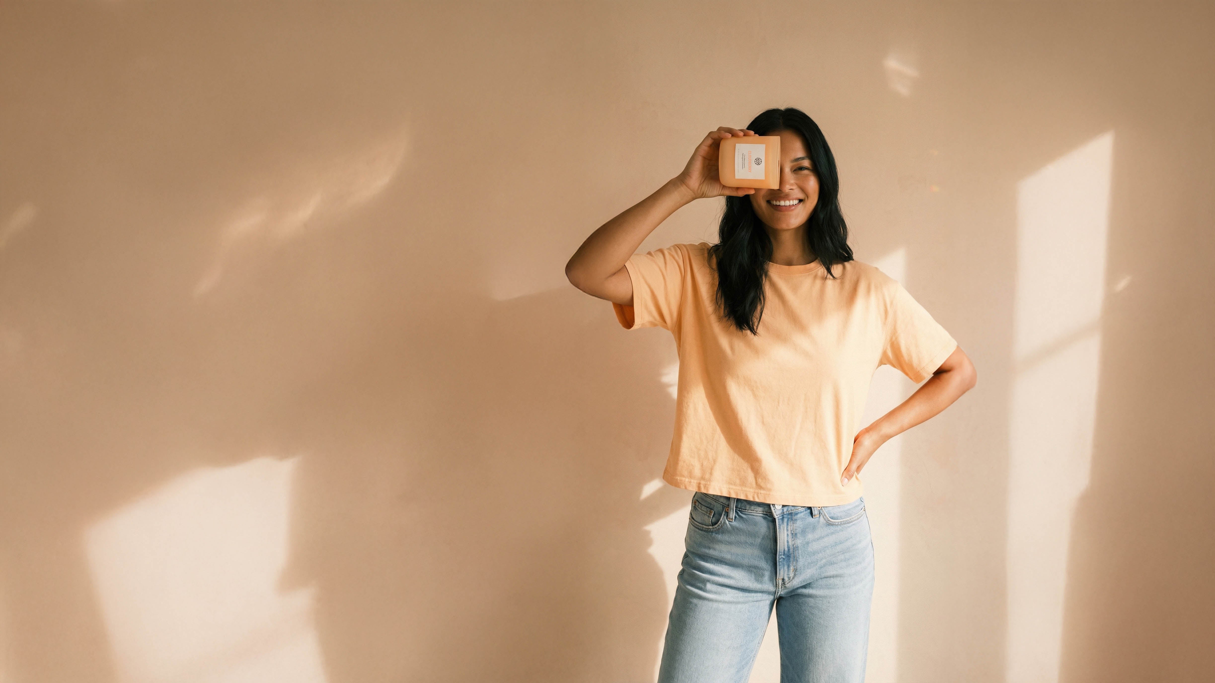 Smiling woman in peach shirt holding AEMBR non-toxic product in sunlit minimal room