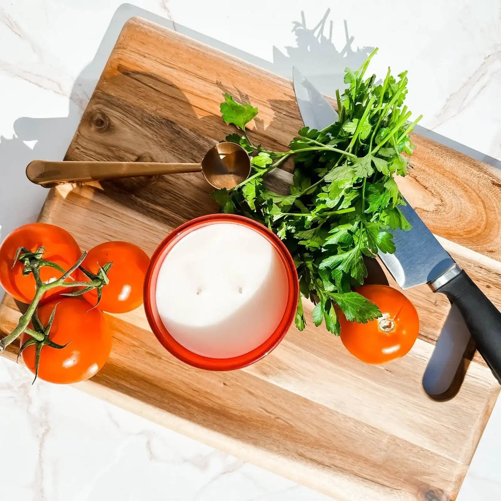 Tomato candle on wood cutting board with tomatoes, parsley, gold spoon, and kitchen knife