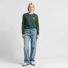 Woman wearing AEMBR dark green long sleeve shirt and light blue jeans, standing on plain background