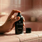 Hand spraying AEMBR ALKYMIST multi-use fragrance bottle 'Love Spell' on pink bathroom tiles
