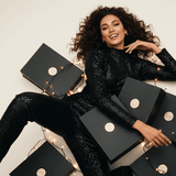 Woman in a black sequin outfit surrounded by black boxes with gold accents on a light background
