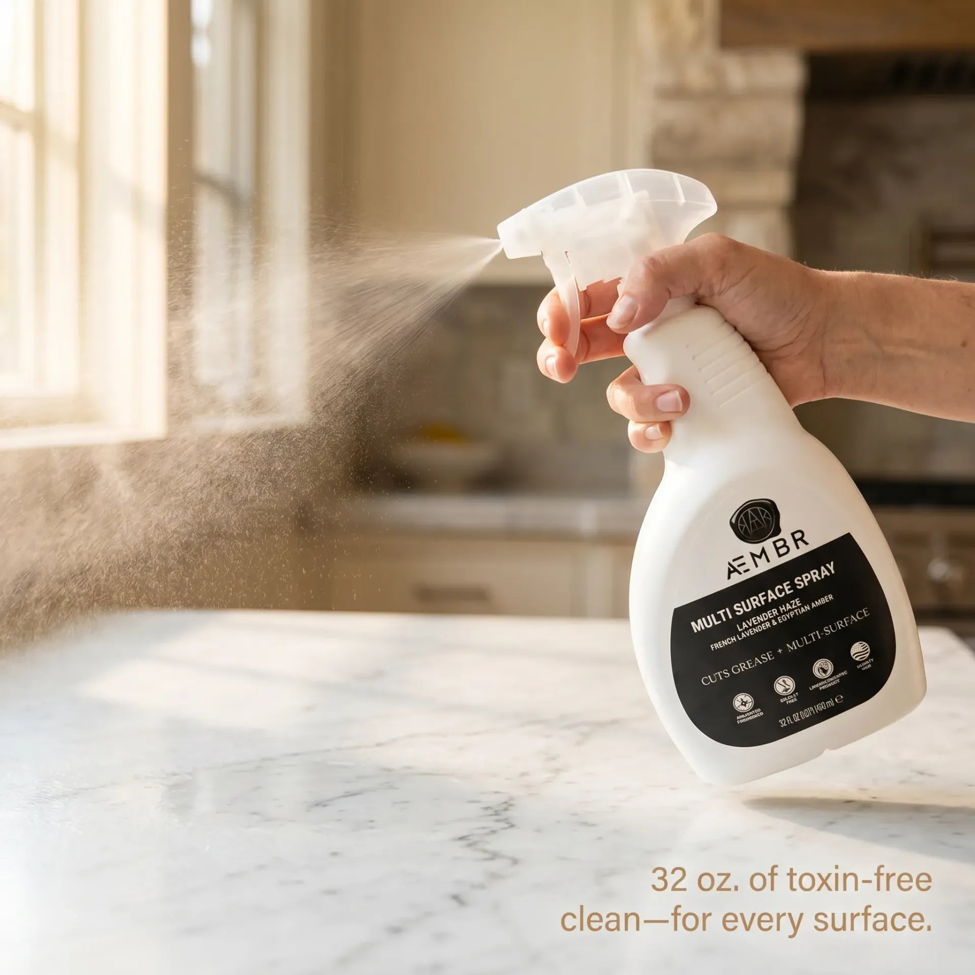 AEMBR Multi Surface Spray in use lifestyle — hand spraying on marble countertop with fine mist, warm kitchen light, 32 oz toxin-free cleaning for every surface