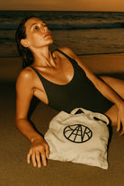 Woman in black swimsuit relaxing on beach with AEMBR branded canvas tote at sunset