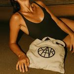AEMBR logo tote bag on sand beside woman in black swimsuit at beach