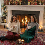 Two women sitting on a rug in front of a fireplace holding cups.