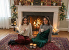 Two women with mugs sit by a lit fireplace, AEMBR candles and holiday decor in cozy living room.