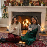 Two women sitting on a rug in front of a fireplace, holding cups, with Christmas decorations around.