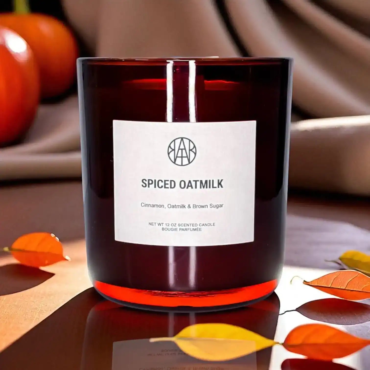 AEMBR Spiced Oatmilk candle in dark glass jar, autumn leaves and pumpkins in cozy home setting.