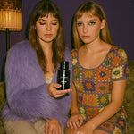 Two women indoors, one in a lavender faux fur jacket holding AEMBR Lavender Haze non-toxic detergent bottle