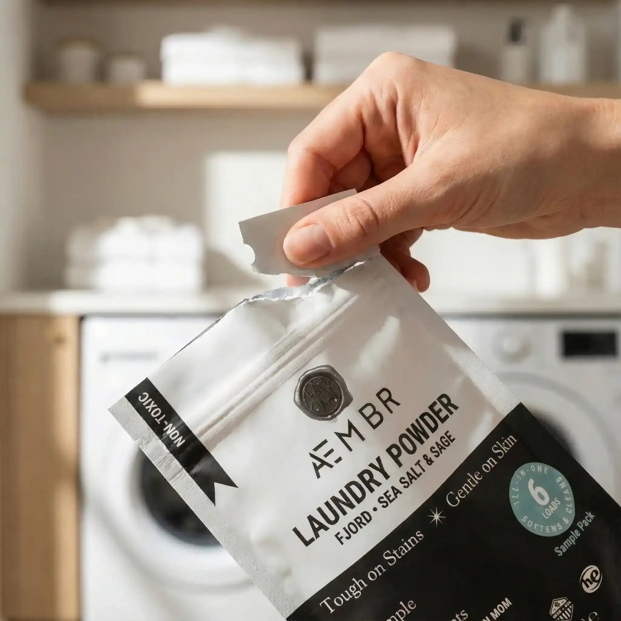 Hand opening AEMBR Laundry Powder trial pack in a modern laundry room, non-toxic detergent.