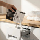 AEMBR non-toxic laundry powder being poured into a washing machine dispenser in a bright laundry room.