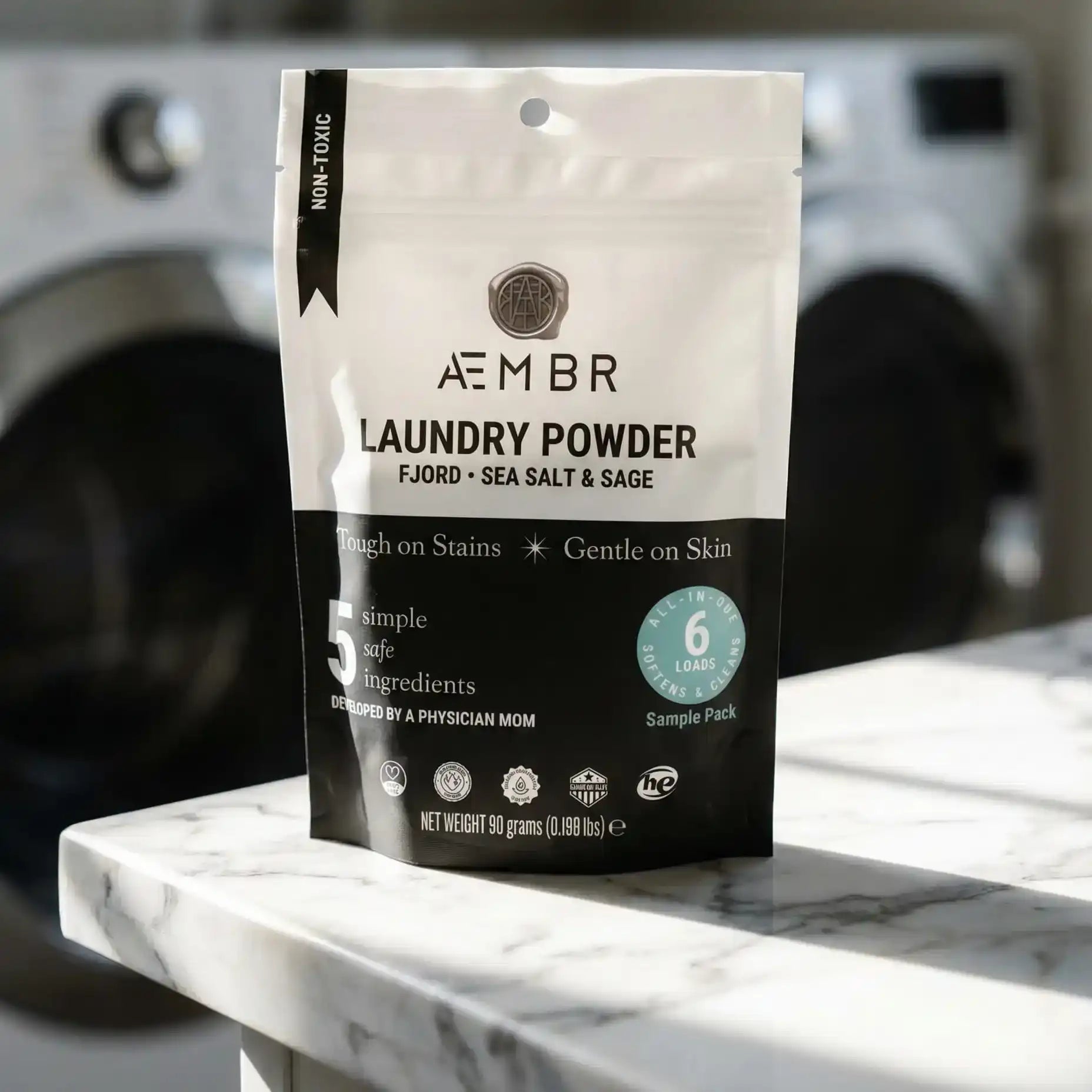 AEMBR Laundry Powder trial pack on marble counter, non-toxic detergent, washing machine background.