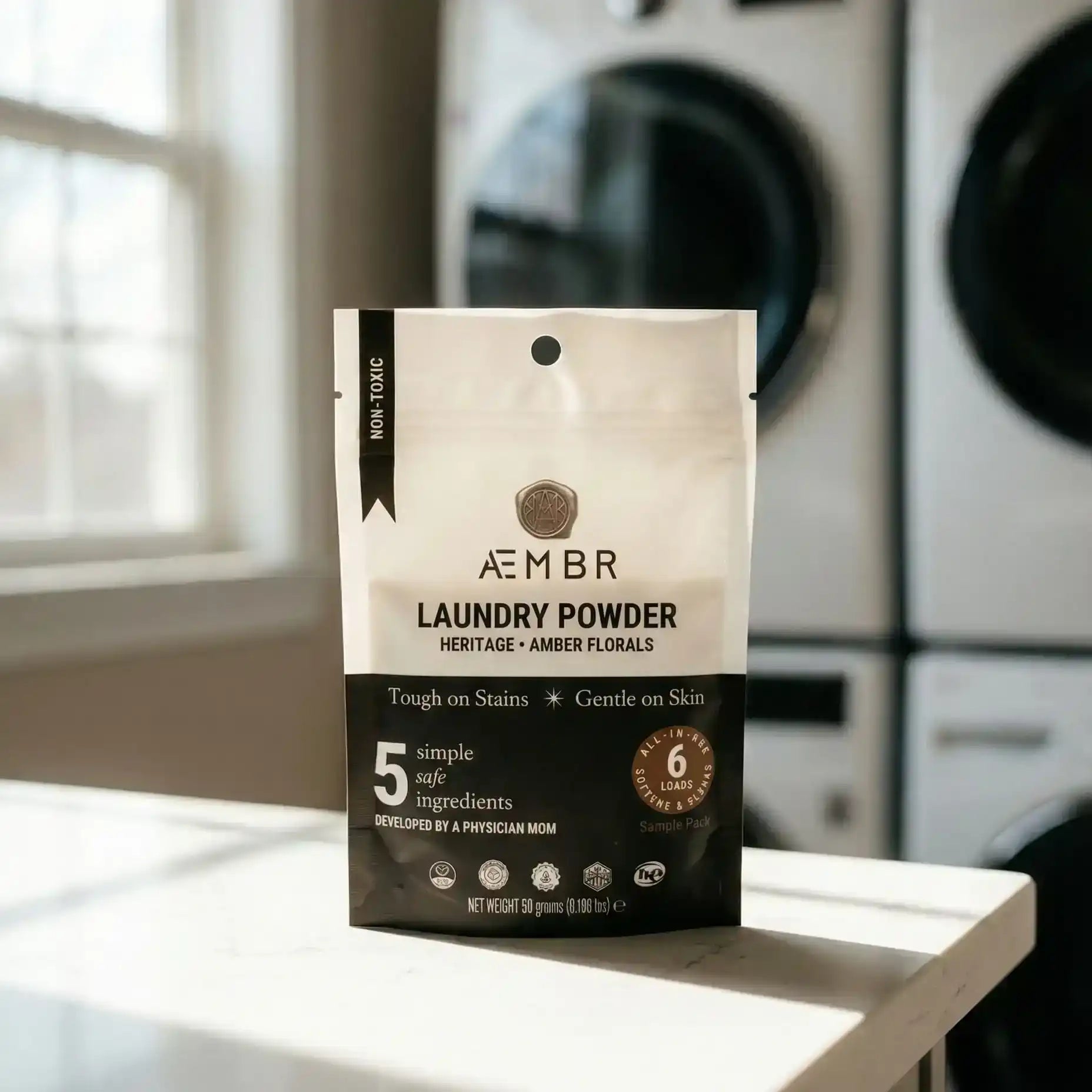 AEMBR non-toxic laundry powder sample pack on a table in a bright laundry room with washer and dryer.