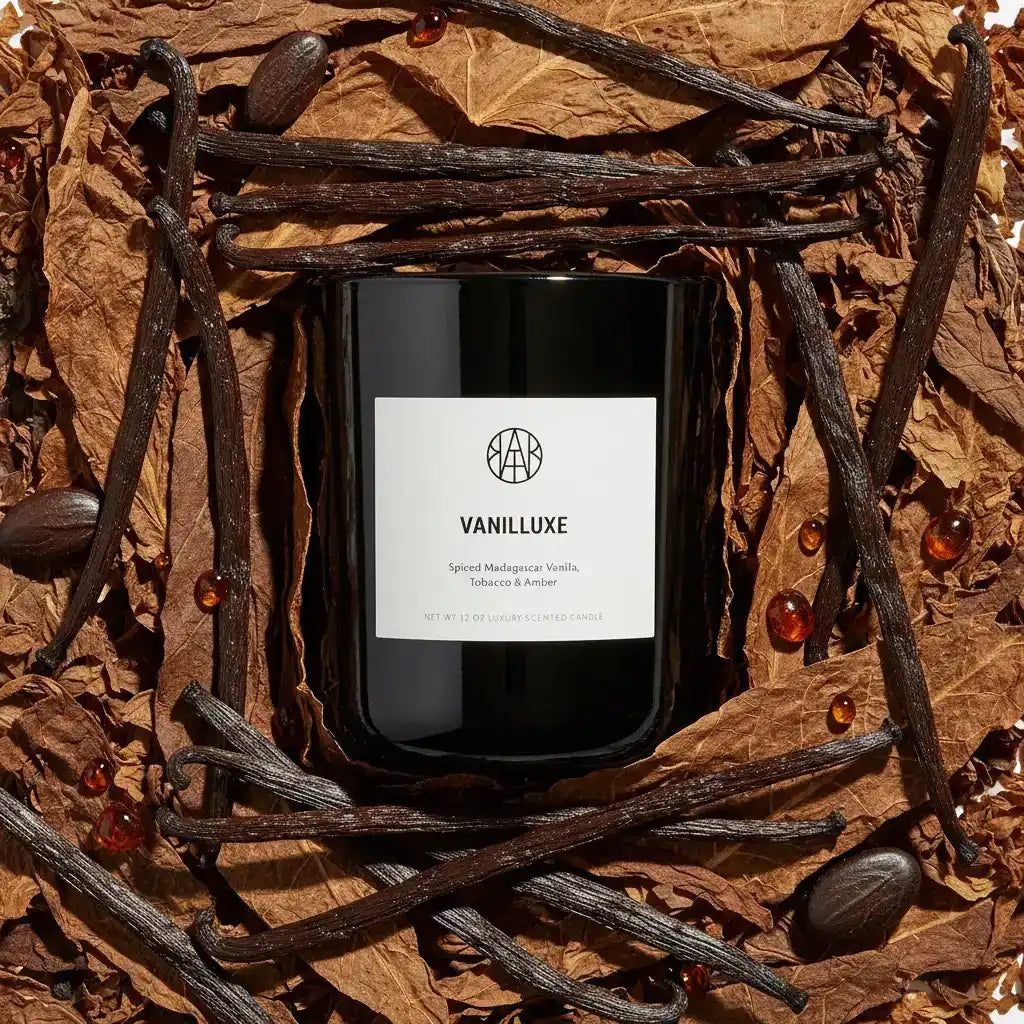 AEMBR VANILLUXE luxury scented candle on dried tobacco leaves, vanilla beans, and amber beads.