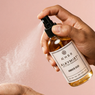 Hand holding a bottle of 'Amber Oud' by AEMBR. ALKYMIST with a spray action on a pink background