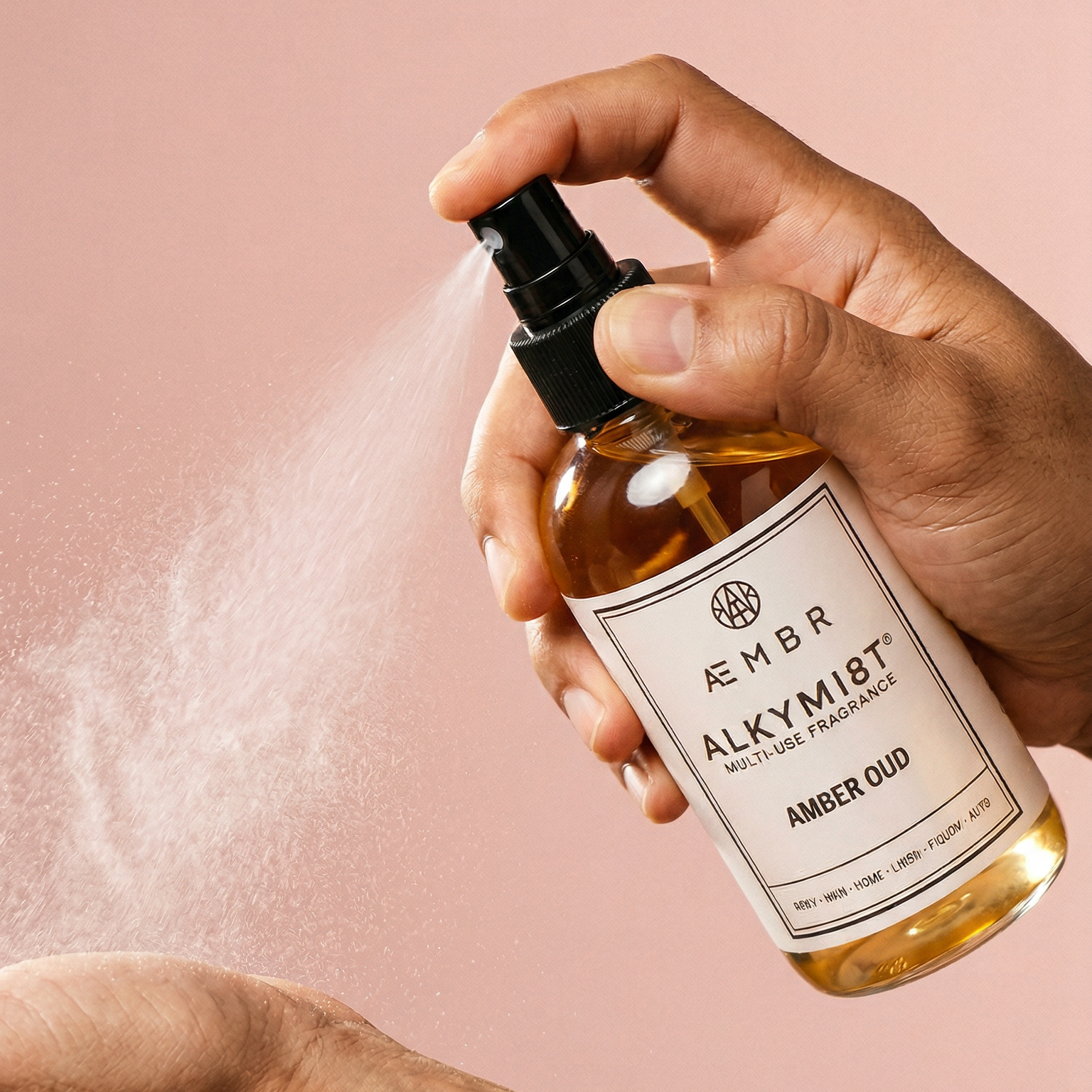 Hand holding a bottle of 'Amber Oud' by AEMBR. ALKYMIST with a spray action on a pink background