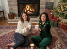 Two women enjoying hot chocolate by a fireplace with a glowing non-toxic candle, holiday decor, gifts, and a Christmas tree.