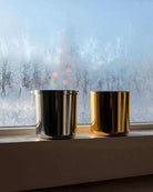 Two metallic non-toxic candles in gold and silver holders on a windowsill with frosted glass.