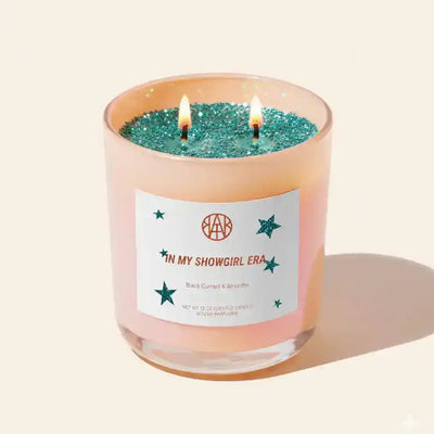 AEMBR In My Showgirl Era candle featuring a sleek glass holder with a lit wick, ideal for creating a cozy ambiance.