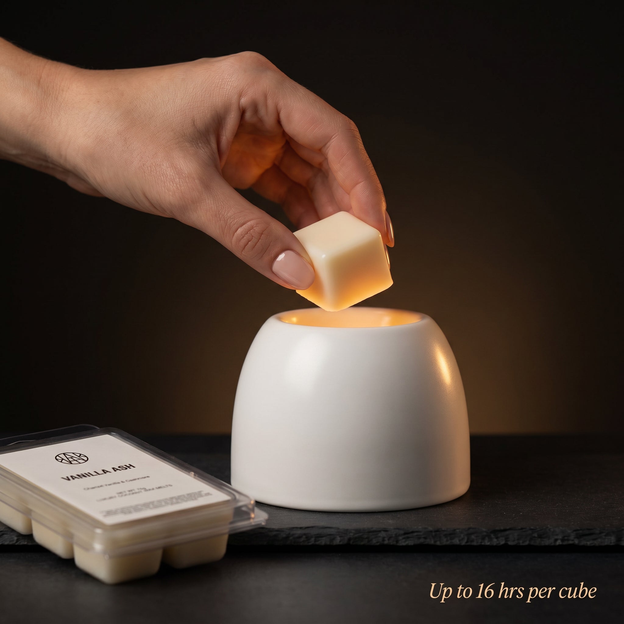 VANILLA ASH Wax Melt in-use lifestyle — hand placing wax cube into white ceramic warmer, amber glow, up to 16 hrs per cube