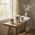 Three white candles with black dots on a wooden table near a window with light curtains.