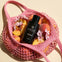 Pink mesh bag with peaches, flowers, and a black bottle labeled 'ALKYMIST LOVE SPELL' on a beige background.