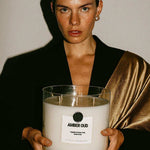Woman holding large AEMBR Amber Oud non-toxic candle, minimalist background.