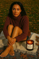 Woman in brown sweater sitting on blanket with AEMBR Pumpkin Teakwood candle outdoors in autumn