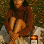 Woman in a brown sweater sitting on a blanket outdoors with a lit AEMBR pumpkin teakwood candle.