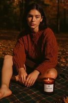Woman in autumn sitting on checkered blanket next to AEMBR Pumpkin Teakwood candle outdoors