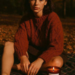Woman in autumn sweater sitting on plaid blanket outdoors with AEMBR pumpkin teakwood candle