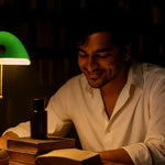 Man reading in cozy library under green lamp with books and a black candle bottle, AEMBR vibe