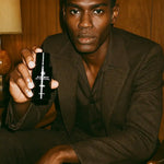 Man in brown suit holding black AEMBR product bottle, seated indoors with wood panel background.