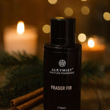 Black fragrance bottle labeled 'Fraser Fir' by Alkymist on a wooden surface with candles and cinnamon sticks in the background.